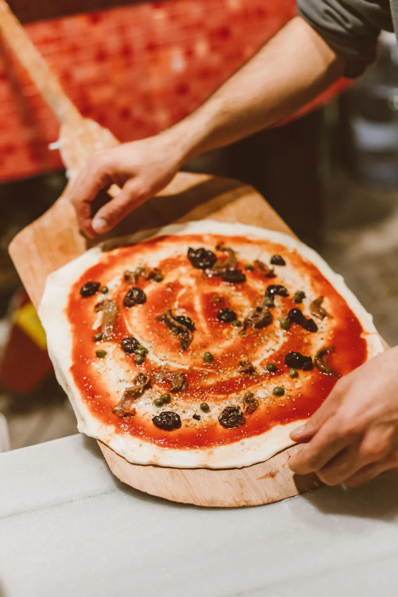 Une main garnit une pizza de sauce tomate et d'olives, sur la pelle en bois